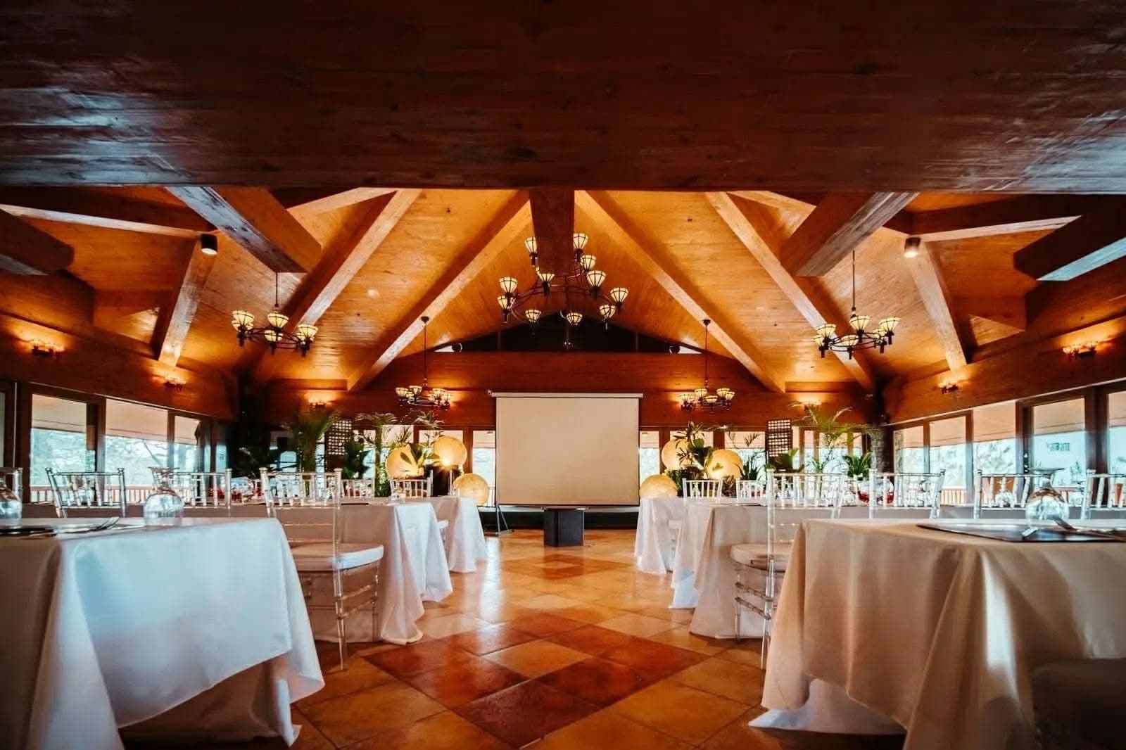 Interior of the 19th Tee at The Forest Lodge, Camp John Hay — wood-beam vaulted ceiling with chandeliers, banquet tables set with white linen, and a wraparound veranda.